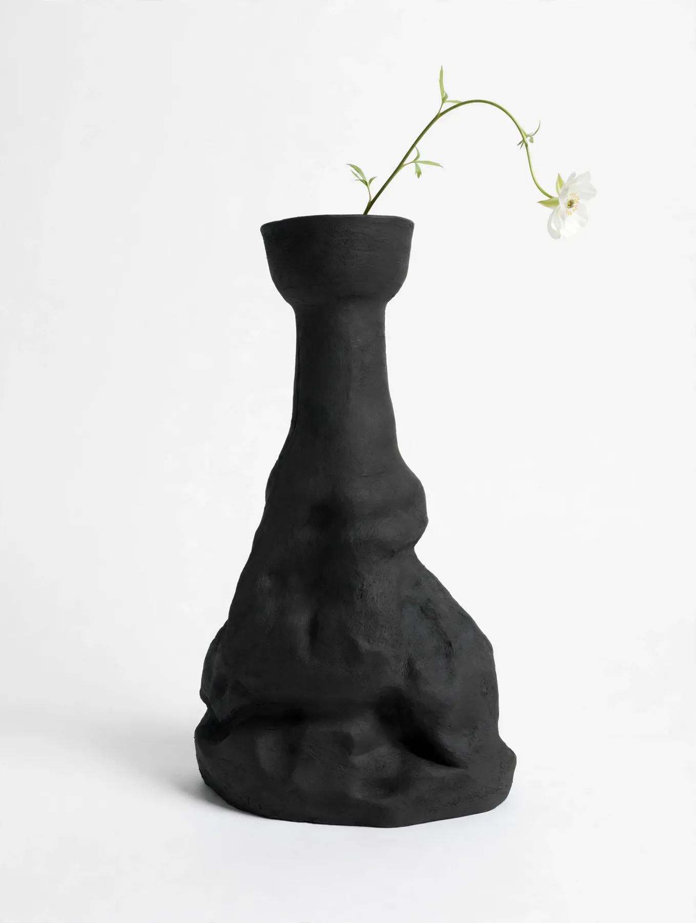 Tall black sculptural piece, massive and rough at the base, holding a single white flower on a long curving stem